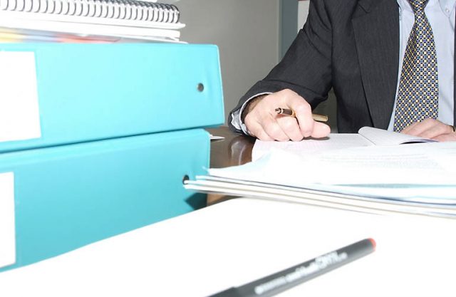 man writing at office desk, stack of folders in front